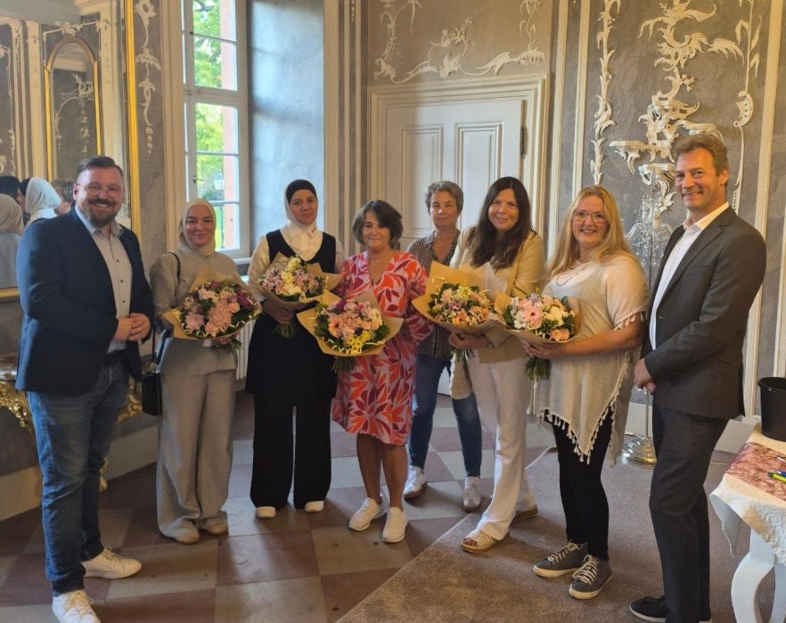 Gruppenfoto in einem historischen Saal mit Stuckverzierungen: mehrere Frauen halten bunte Blumensträuße, links und rechts von ihnen stehen zwei Männer.