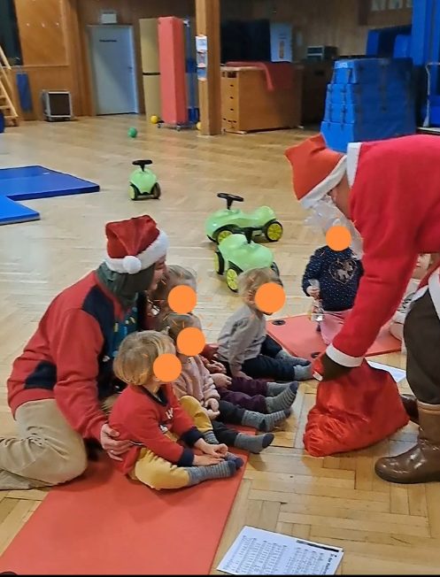 Kleinkinder sitzen mit einer Erwachsenen auf roten Matten in einer Turnhalle. Ein Nikolaus holt Geschenke aus einem roten Sack, im Hintergrund stehen Rutschautos. Die Gesichter der Kinder sind verdeckt.