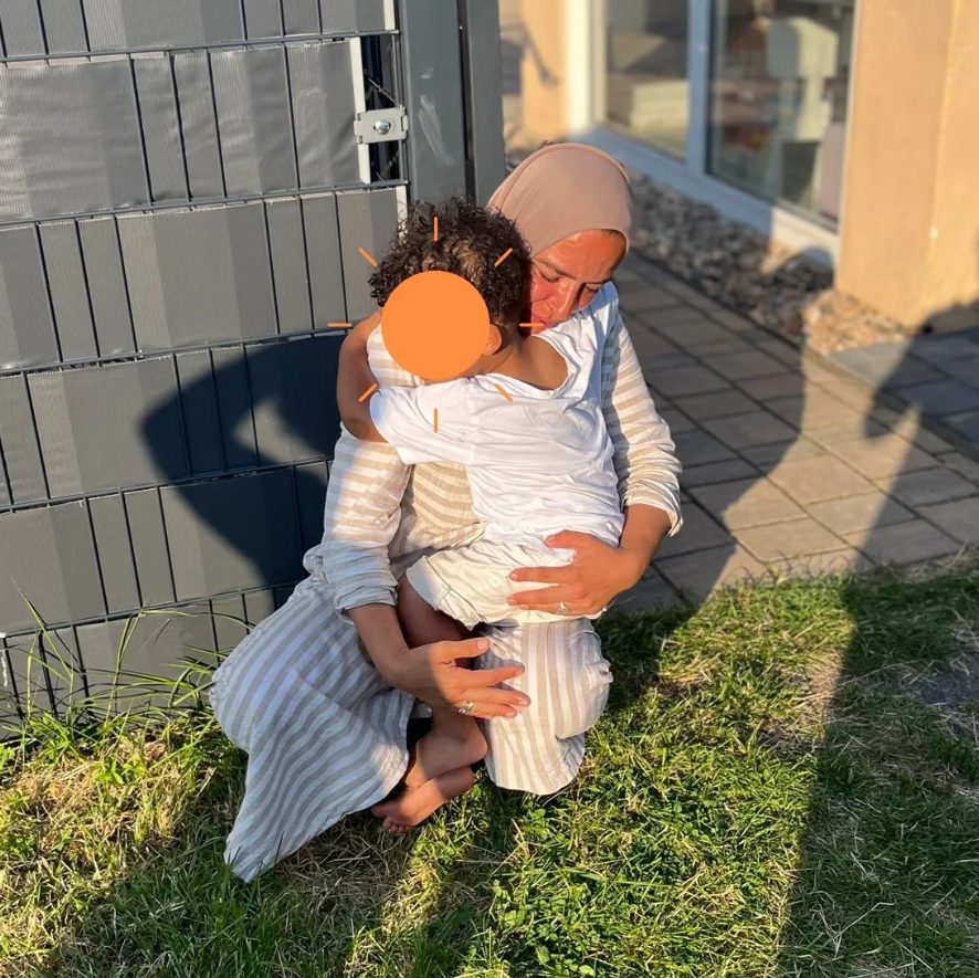 Die Tagesmutter kniet in der Abendsonne auf dem Rasen und hält ein kleines Kind fest im Arm. Das Gesicht des Kindes ist verdeckt.